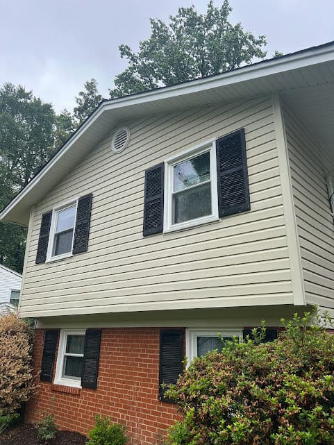Two-story house with beige siding, black shutters, and a brick foundation, surrounded by greenery.