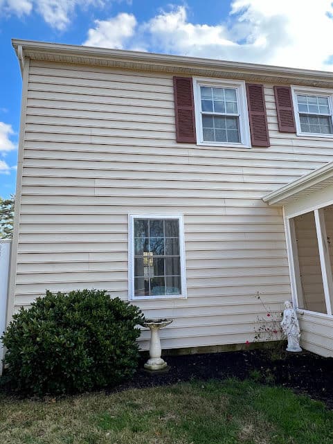 Two-story beige house with brown shutters and garden statue by the window on a sunny day.