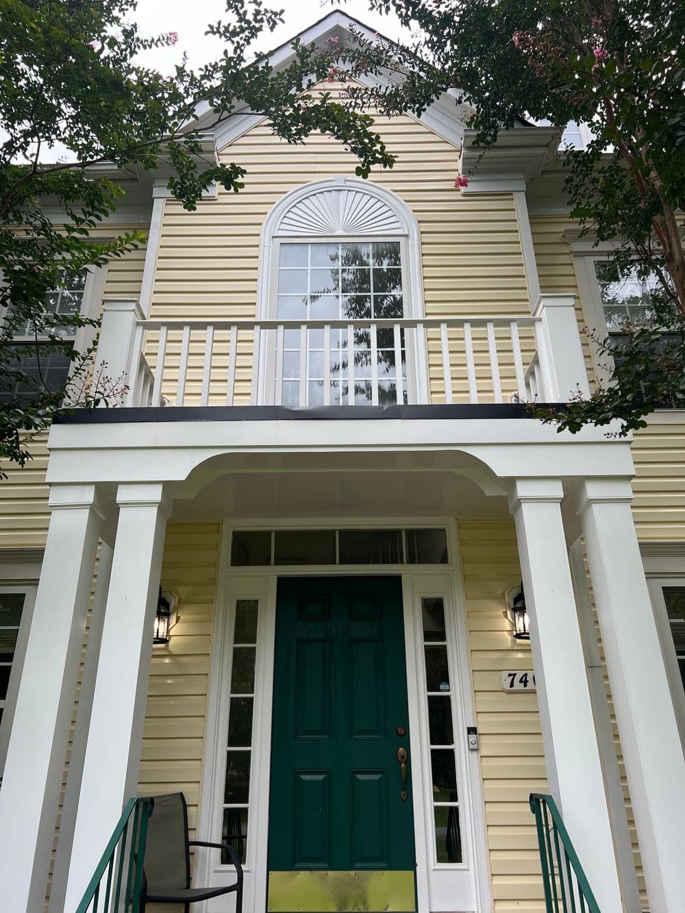 Charming yellow house with green door and balcony, surrounded by trees and shrubs.