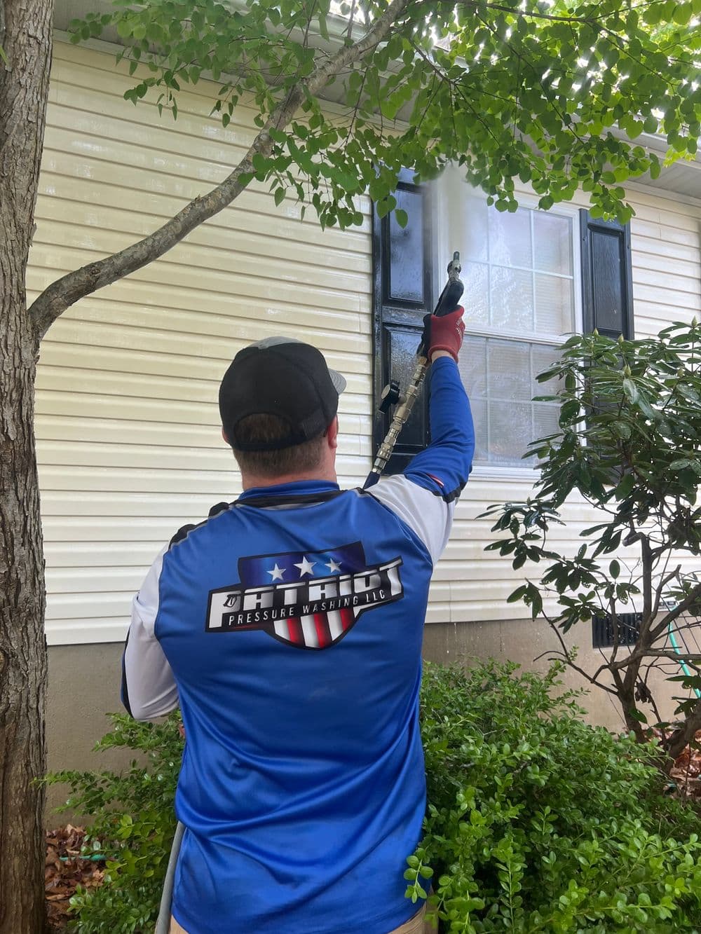 Person using a pressure washer on a house window behind greenery.