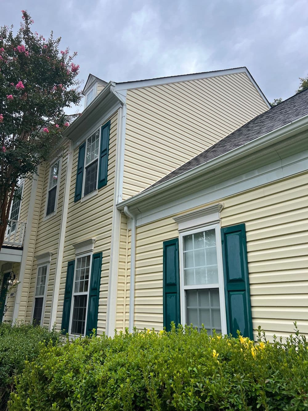 Yellow house with green shutters and nearby flowering bush under a cloudy sky.