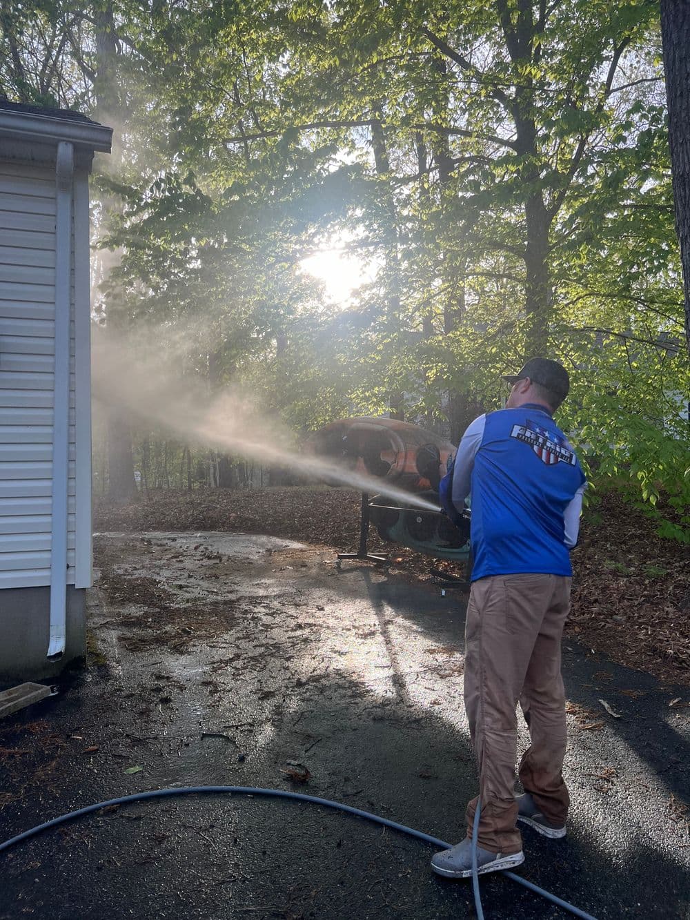 Man using a pressure washer to clean a driveway surrounded by trees and sunlight.